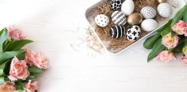 Üst manzara, altın ve siyah ve beyaz sanat, yeni Paskalya tarzı yumurtalar, beyaz bir masadaki pembe püsküllü lalelerin yanında, bir tepside samanlıkta yatıyor. Konforlu İskandinav bayramı dekoru.