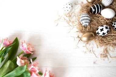 Üst manzara, altın ve siyah ve beyaz sanat, yeni Paskalya tarzı yumurtalar, beyaz bir masadaki pembe püsküllü lalelerin yanında, bir tepside samanlıkta yatıyor. Konforlu İskandinav bayramı dekoru.