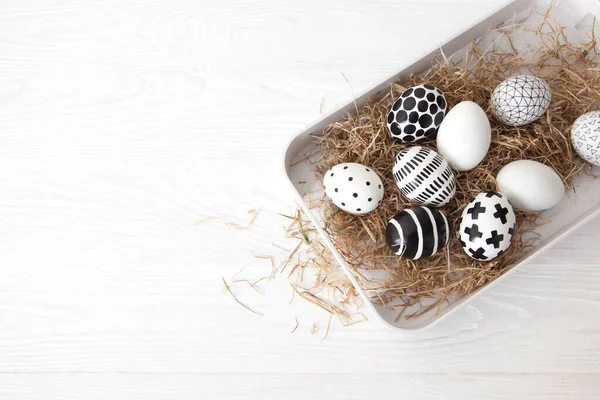 Close-up of golden and black and white minimalist painted easter eggs lie in the hay on a white tray with sides on a white table. Stylish modern rustic easter concept. Copyspace