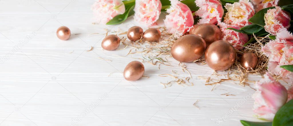 Golden different sizes eggs lie on a white wooden table next to hay and beautiful pink tulips. The concept of spring holidays and Easter in the European style. Copyspace