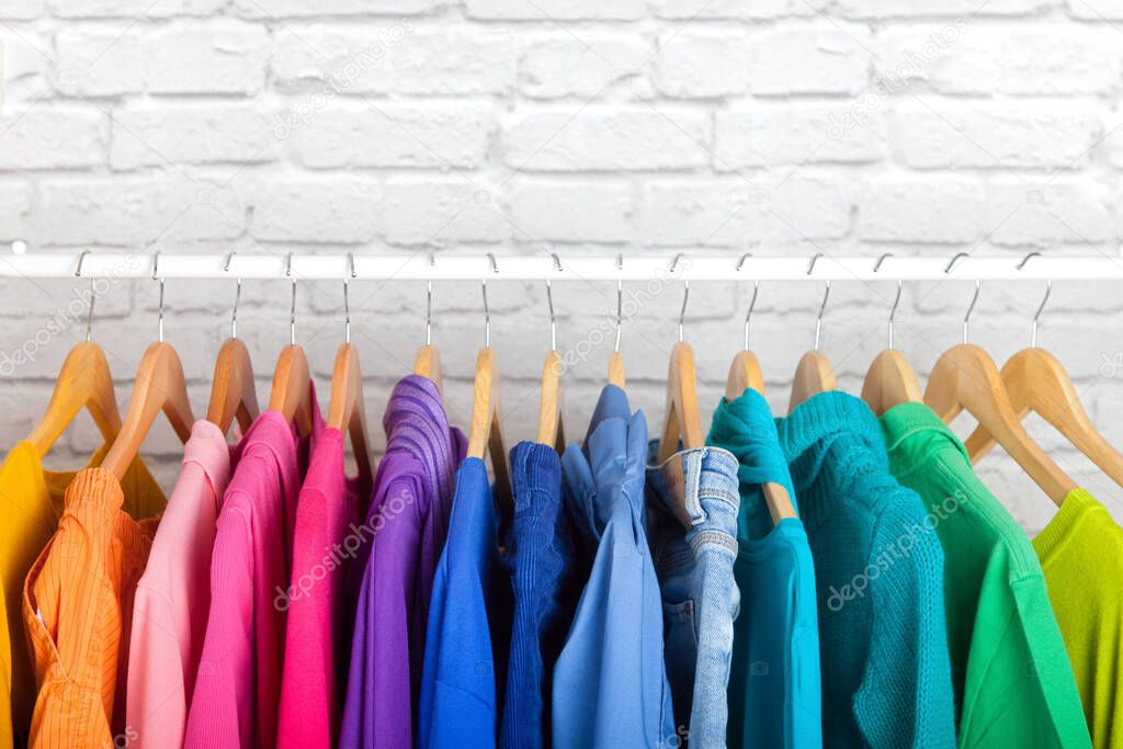 Women's wardrobe sweatshirts shirts and blouses hang on wooden shoulders in the order of all the colors of the rainbow against white brick wall. Concept update in the wardrobe. Advertising space