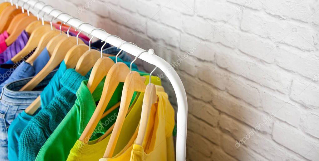 Close-up clothes of all colors of the rainbow hanging on wooden hangers on an iron floor hanger against a blurry brick wall. Concept of an orderly wardrobe and stylish clothes