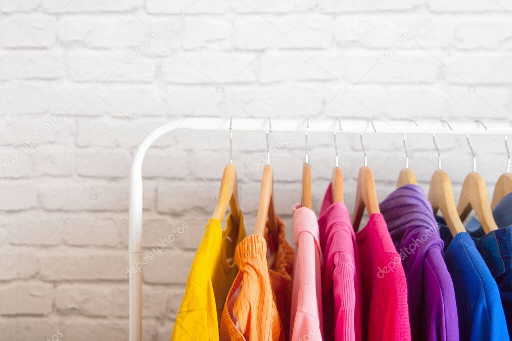 Close-up clothes of all colors of the rainbow hanging on wooden hangers on an iron floor hanger against a blurry brick wall. Concept of an orderly wardrobe and stylish clothes