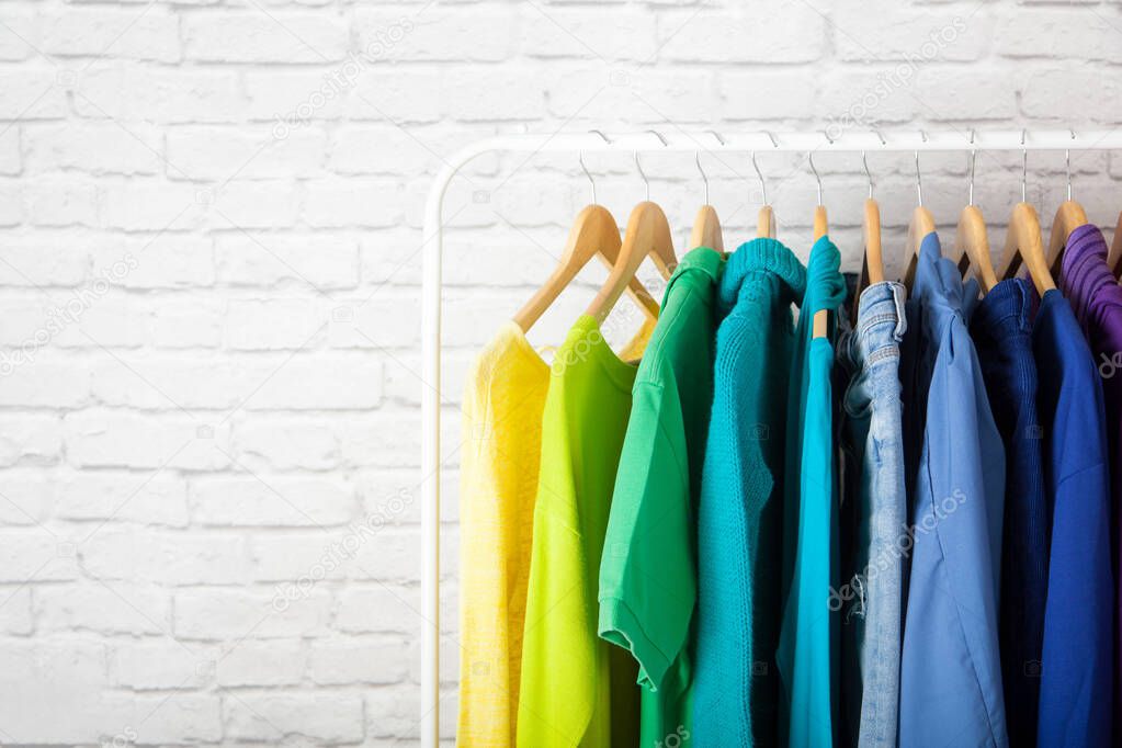 Women's wardrobe sweatshirts shirts and blouses hang on wooden shoulders in the order of all the colors of the rainbow against white brick wall. Concept update in the wardrobe. Advertising space