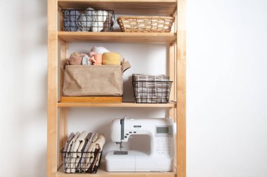 Farklı dikiş malzemeleri ve iplikler sepetlerde yatar ve beyaz bir arka planda dikiş makinesi olan ahşap bir rafta dururlar. Terzi ve nakış işyeri kavramı. Kopyalama alanı