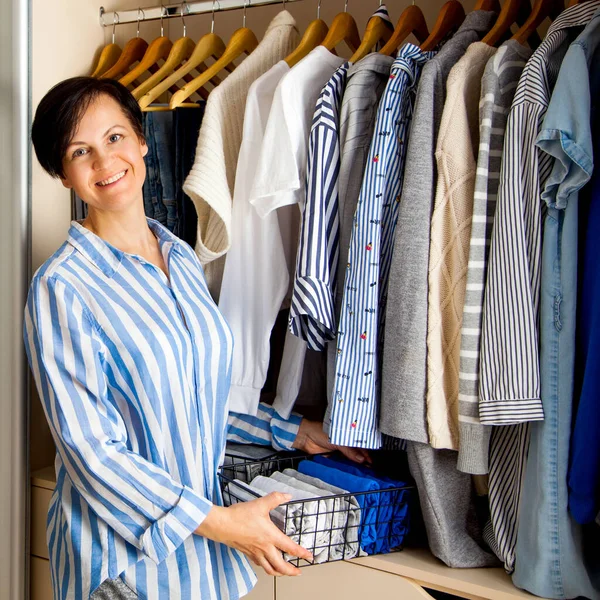 Pretty positive caucasian young woman housewife is holding a container with things of blue shades against the background of her wardrobe with perfect order. Lady fly concept