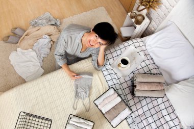 Şık bir yatak odasında bir fincan kahveyle yatağın yanında otururken gardırobundaki eşyaları toplayan neşeli, güzel esmer kadın manzarası. Bahar temizliği kavramı