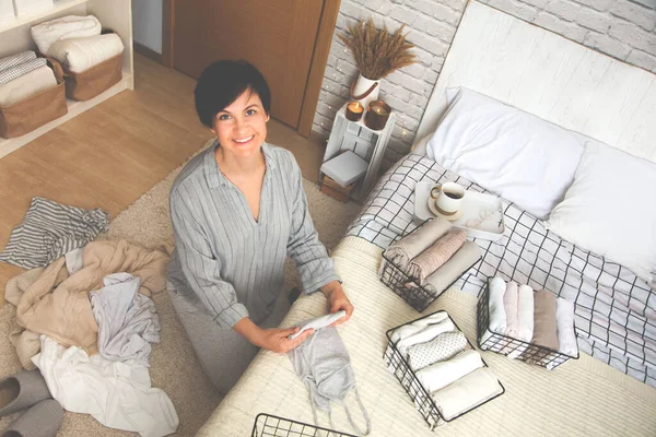 Güzel, genç, esmer bir kadının rahat yatak odasında otururken bir şeyleri örgü metal kaplar üzerinde katlamasının en güzel görüntüsü. Haftalık temizlik konsepti