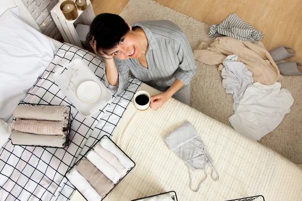 Top view of cheerful beautiful young brunette woman tidying up things in her wardrobe while sitting on floor near bed in stylish bedroom with cup of coffee. Spring tidy concept