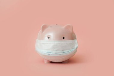 Tıbbi maskeli pembe domuzcuk kumbarası soluk pembe arka planda duruyor. Donmuş banka hesapları ve mevduat kavramı ve zor zamanlar için birikmiş para. reklam alanı