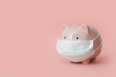 Tıbbi maskeli pembe domuzcuk kumbarası soluk pembe arka planda duruyor. Donmuş banka hesapları ve mevduat kavramı ve zor zamanlar için birikmiş para. reklam alanı