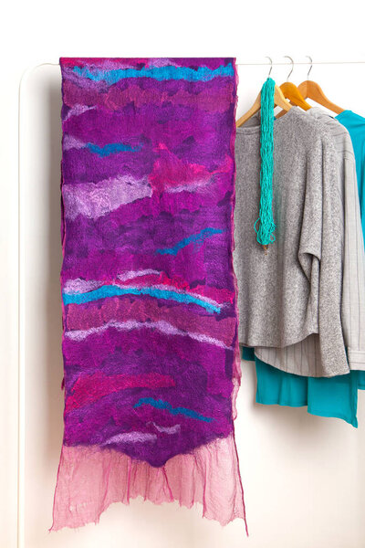 Beautiful large felted purple silk woolen homemade scarf hanging on white hanger next to gray blue shirts on white background in studio. Spring wardrobe concept