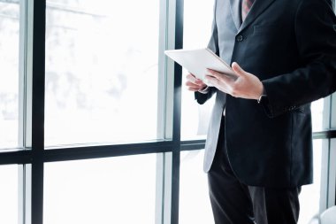 Ofis penceresinin yanında tabletle ayakta duran genç işadamı, iş konsepti