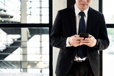 Ofis penceresinin yanında akıllı telefon la ayakta duran genç işadamı, iş konsepti