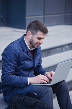 Bina merdivenlerde otururken dizüstü bilgisayarda yazarak mutlu gülümseyen iyi giyimli iş adamı 