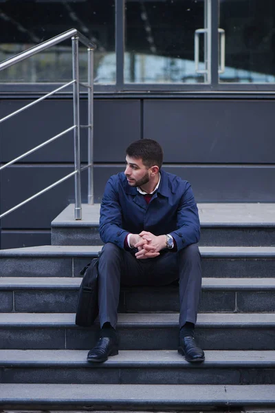 upset well-dressed businessman looking aside while sitting on stairs of building