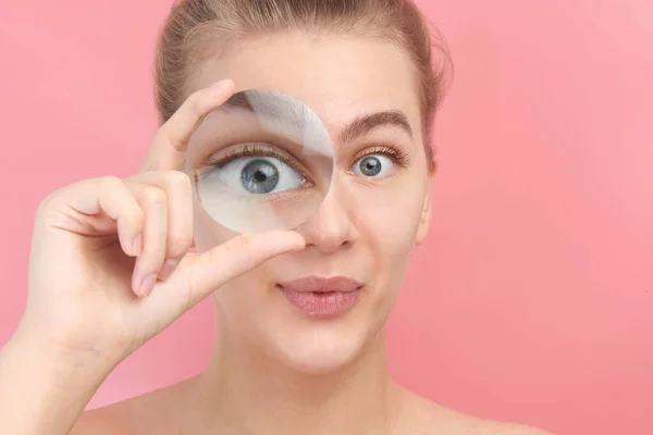 Young beautiful woman looking at the camera through magnifier with  funny surprised expression, closeup isolated on pink. Enlarged eye, vision concept