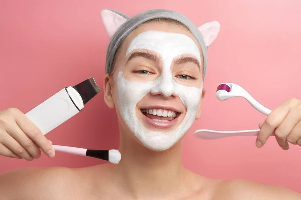 Close-up of beautiful young girl with white mask on face and Korean cat ears headband smiling and demonstrates various home and salon skin care products in pink studio