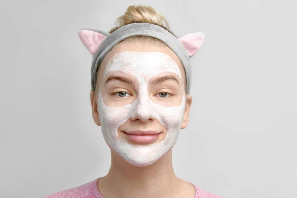 Cute young kawaii girl with cosmetic white mask on her face and korean cat ears headband portrait in white studio, skin care