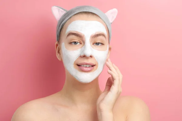 Young woman with cat ears applied white cleansing mask to her face, touches and enjoys the smoothness and softness of skin, isolated in pink studio