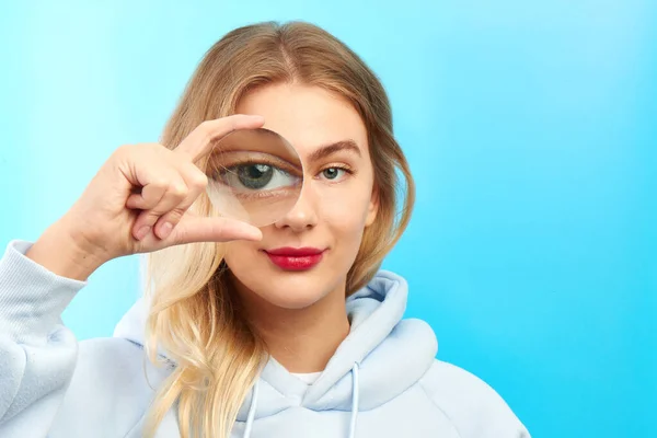 Young spy woman looking at camera through magnifier, big eye is watching you closeup isolated on blue. Enlarged eyes, optical illusion, search