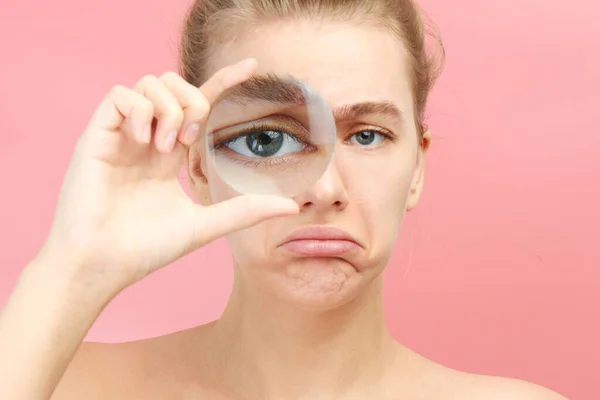 Young upset woman looking at the camera through magnifier big eye closeup isolated on pink. Bad vision, search concept
