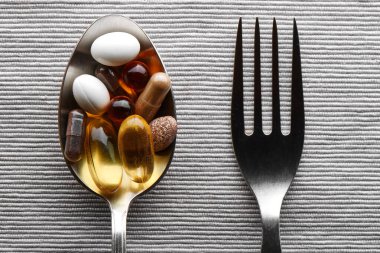 Gri kumaş arka planında çeşitli besin takviyeleri, tabletler ve vitaminlerle dolu bir yemek kaşığı ve çatalın yakın çekimi