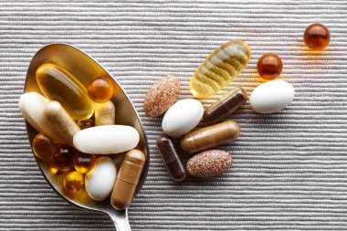 Gri kumaş arka planında çeşitli besin takviyeleri, tabletler ve vitaminlerle dolu yemek kaşığı.
