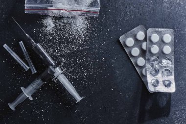 Uyuşturucu bağımlılığı kavramı, bağımlılık. Dağınık kokain veya eroin tozu, şırıngalar ve siyah arka planda haplarla su toplaması.
