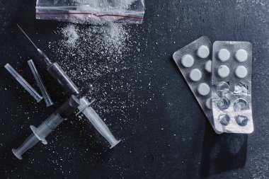 The concept of drug dependence, addiction. Scattered cocaine or heroin powder, syringes and blister with pills on black background top view clipart