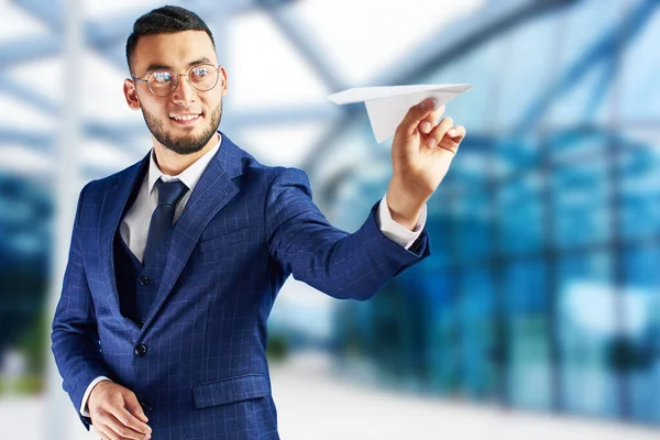 Smiling Asian Kazakh businessman in suit and glasses holds paper airplane on blue blurred background. Small business development concept, project launch, team training, start-ups, global strategy