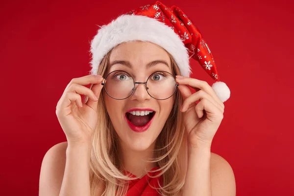 Surprised woman in glasses and christmas santa claus hat with open mouth and bulging eyes looks camera, sees something incredible amazing isolated on red