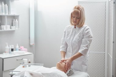 Profesyonel güzellik uzmanı, beyaz güzellik salonunda genç bir kadına yüz ve boyun bölgesi için elle masaj yapıyor. Kozmetoloji, yüz bakımı prosedürü