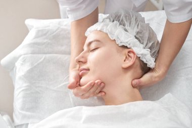 Profesyonel güzellik uzmanı, beyaz güzellik salonunda genç bir kadına yüz ve boyun bölgesi için elle masaj yapıyor. Kozmetoloji, yüz bakımı prosedürü
