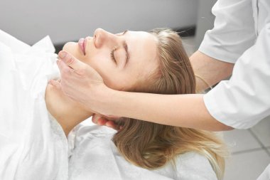 Profesyonel güzellik uzmanı, beyaz güzellik salonunda genç bir kadına yüz ve boyun bölgesi için elle masaj yapıyor. Kozmetoloji, yüz bakımı prosedürü