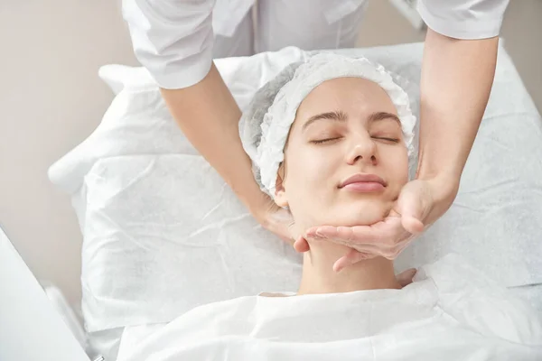 Professional beautician gives young woman manual massage of face and cervical collar zone in white beauty parlor. Cosmetology, face care procedure