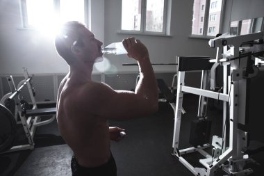 Çıplak gövdeli güçlü atletik adam spor salonunda elektrolitlerle su içer. Vahşi vücut geliştirici egzersizi, spor beslenme konsepti.