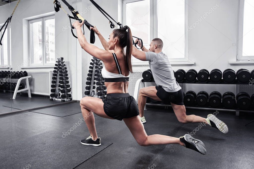 Fitness sexy brunette woman dressed top and shorts pumps arms and legs muscles with trainer in gym, personal workout program, TRX straps training