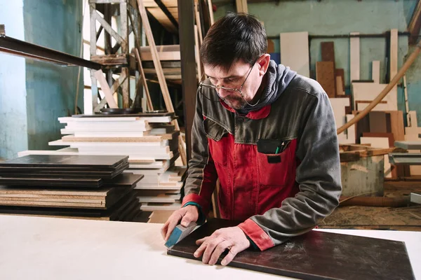 Process of production and manufacture of wooden furniture in furniture factory. Worker carpenter man in overalls processes wood on special equipment