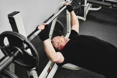 Yetişkin, gri saçlı adam spor salonunda spor aletleriyle antrenman yapıyor, bacak ve kol kaslarını çalıştırıyor, kilo veriyor. Yaşlılıkta sağlıklı yaşam biçimi kavramı