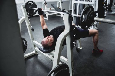 Yetişkin, gri saçlı adam spor salonunda spor aletleriyle antrenman yapıyor, bacak ve kol kaslarını çalıştırıyor, kilo veriyor. Yaşlılıkta sağlıklı yaşam biçimi kavramı