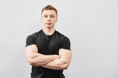 Siyah tişörtlü yakışıklı genç sporcunun portresi beyaz arka planda kolları çaprazlanmış şekilde gülümsüyor.