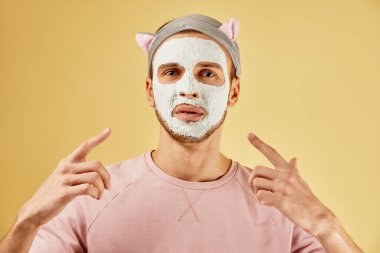 Beyaz kozmetik maskeli sakallı komik bir adamın portresi sarı arka planda izole edilmiş bir parmak izi gösteriyor.