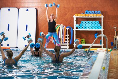 Genç kızlar ve orta yaşlı kadınlar su aerobiği yapıyorlar. Spor hocasıyla havuzda dambıllar takıyorlar. Suda kilo vermek için spor yapıyorlar.