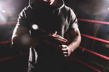 Closeup portrait professional boxer in a hood wraps the palms of his hands, puts on gloves in the ring. Dark colors, no face clipart