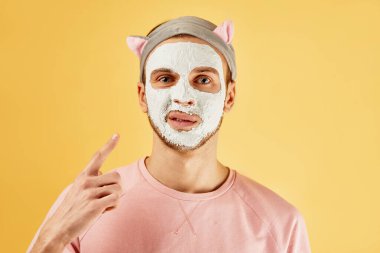 Beyaz kozmetik maskeli sakallı komik bir adamın portresi sarı arka planda izole edilmiş bir parmak izi gösteriyor.