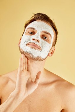 Beyaz maskeli yakışıklı sakallı genç adamın portresi sarı arka planda, erkek cilt bakımı kavramına izole edilmiş bir şekilde stüdyoda yüzüne dokunuyor.