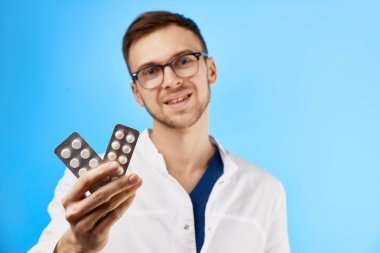 Gözlüklü ve beyaz önlüklü nazik doktor haplardan su toplamış ve mavi arka planda izole edilmiş kameraya gülümsüyor. Dokunulmazlığı ilaçlarla destekle, virüse ve gribe karşı koruma