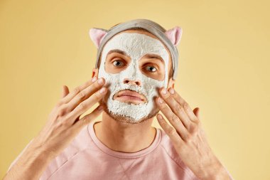 Yüzüne beyaz maske takan sakallı komik bir adamın portresi, kameraya sarı arka planda izole edilmiş bir ayna gibi bakıyor, erkek derisi konsepti.