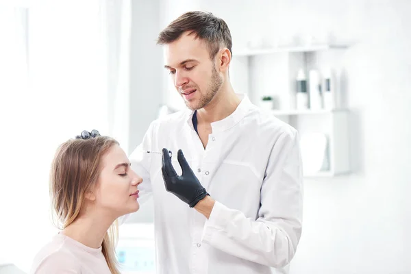 美容師男性は若い女性患者に美容注射を与えます ハンサムな皮膚科医は額のしわにボツリヌス毒素を置きます 美容室での顔充填 — ストック写真